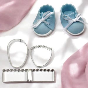 Puede incluir: Dos zapatos de bebé azul claro con cordones blancos y suela blanca. También se muestran tres cortadores de galletas de metal plateado: un óvalo, una forma de pie y un rectángulo festoneado. Dispuestos sobre una superficie blanca con tela rosa.