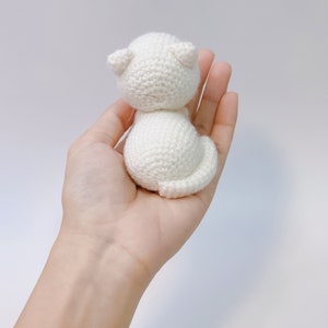May include: White crocheted cat amigurumi toy. The cat is sitting up with its tail curled around its body.