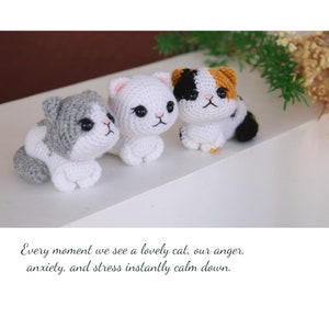 May include: Three handmade crochet cat figurines in gray and white, white, and calico patterns. The cats are sitting on a white surface. The text at the bottom reads: Every moment we see a lovely cat, our anger, anxiety, and stress instantly calm down.