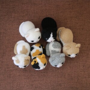 May include: A collection of handmade crochet cat toys in various colors, including white, black, gray, and tan, with detailed facial features. The toys are arranged on a brown surface.