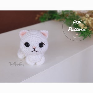 May include: A white crocheted cat amigurumi with pink ears and black eyes. The cat is sitting on a white surface. The text "PDF Pattern ENGLISH" is visible in the background.