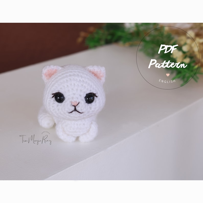 May include: A white crocheted cat amigurumi with pink ears and black eyes. The cat is sitting on a white surface. The text "PDF Pattern ENGLISH" is visible in the background.
