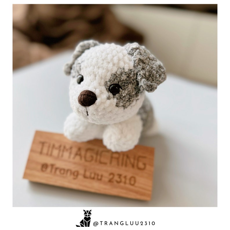 May include: A crocheted white and gray dog toy with brown eyes and a brown nose. The toy is sitting on a wooden sign that says "Timmagicring @frang_luu_2310".