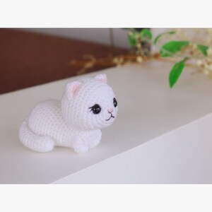 May include: White crocheted cat plush toy with black eyes and pink nose. The cat is lying on its side with its head turned to the right.
