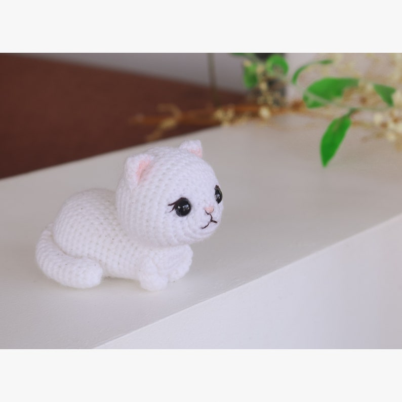 May include: White crocheted cat plush toy with black eyes and pink nose. The cat is lying on its side with its head turned to the right.