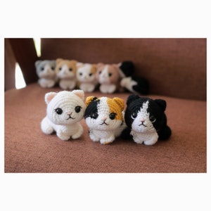 May include: Three crocheted cat plushies in white, black, and orange with black eyes and pink noses. The plushies are sitting on a brown surface.