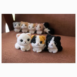 May include: A collection of handmade crochet cat figurines in various colors, including white, calico, and black and white. The small cat toys are arranged on a brown surface, with several more in the background. The cats have detailed features.