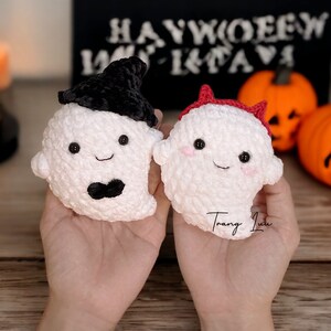 Op de afbeelding: Twee gehaakte spook knuffels, een met een zwarte heksenhoed en de andere met een rood duivelshoorn hoofdband. De knuffels worden in de handen van een persoon gehouden tegen een houten achtergrond met pompoenen en kaarsen.
