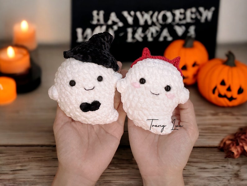 Op de afbeelding: Twee gehaakte spook knuffels, een met een zwarte heksenhoed en de andere met een rood duivelshoorn hoofdband. De knuffels worden in de handen van een persoon gehouden tegen een houten achtergrond met pompoenen en kaarsen.