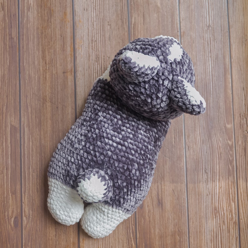 Op de afbeelding: Een grijs en wit gehaakt knuffeldier in de vorm van een hond. De hond ligt op zijn zij met zijn hoofd naar de zijkant gedraaid. De hond heeft een pluche textuur en een witte vlek op zijn borst.