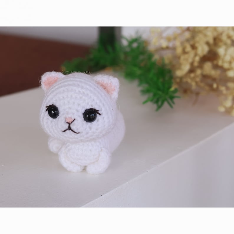 May include: White crocheted cat plush toy with black eyes and pink ears. The cat is sitting on a white surface.