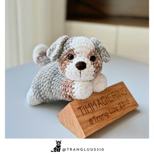 May include: A crocheted stuffed dog with gray, brown, and white fur. The dog is lying down with its head resting on a wooden sign that says "Timmagicring @Trang Luu 2310".