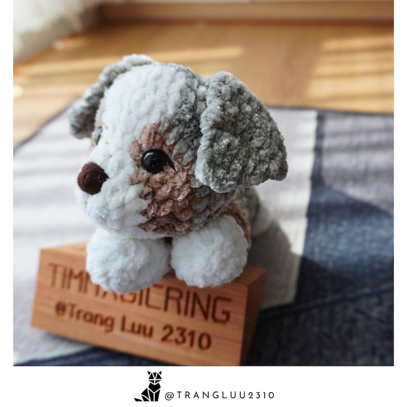 May include: A crocheted stuffed dog toy with white, brown, and gray fur. The dog is sitting on a wooden block with the text "TIMAGILERING @Trang Luu 2310" on it.