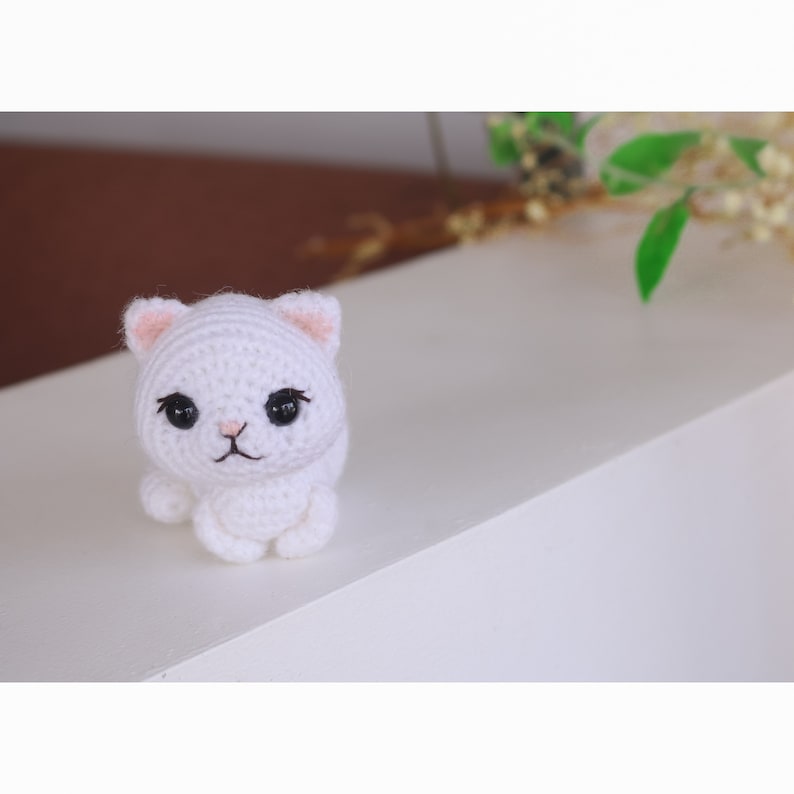 May include: A white crocheted cat with black eyes and pink ears. The cat is sitting on a white surface.
