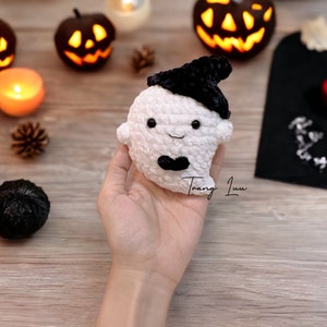 Op de afbeelding: Een witte gehaakte geest met zwarte ogen en een zwarte hoed. De geest houdt een zwarte strik vast. De geest zit op een hand voor een houten achtergrond met Halloween-decoraties.