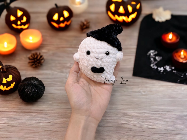 Op de afbeelding: Een witte gehaakte geest met zwarte ogen en een zwarte hoed. De geest houdt een zwarte strik vast. De geest zit op een hand voor een houten achtergrond met Halloween-decoraties.