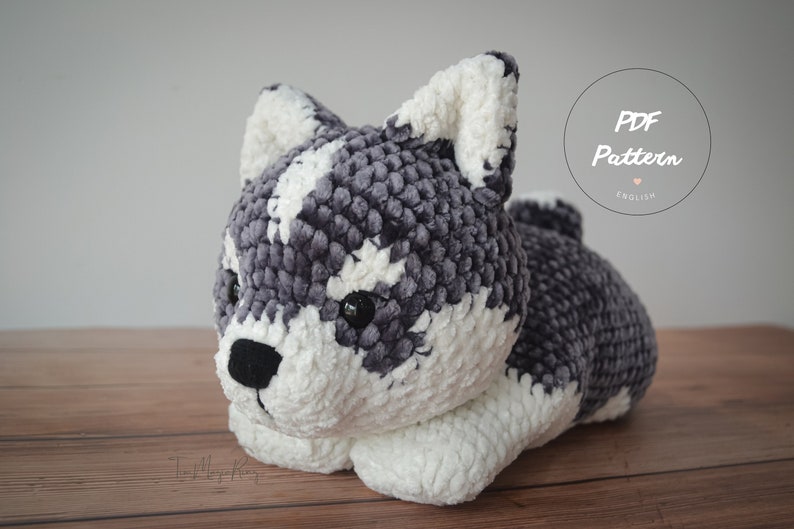 Op de afbeelding: Gehaakt pluche husky hondenspeelgoed in grijs en wit met zwarte ogen en neus. Het speelgoed ligt met zijn kop op zijn poten. Het speelgoed ligt op een houten oppervlak. PDF Pattern English.