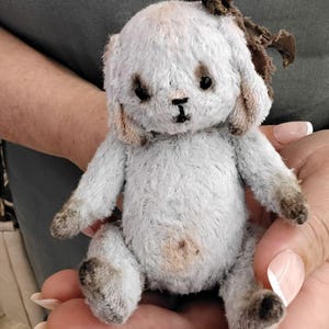 May include: A small, gray and white plush bunny with a brown fabric bow on its head. The bunny is sitting in a person's hand.