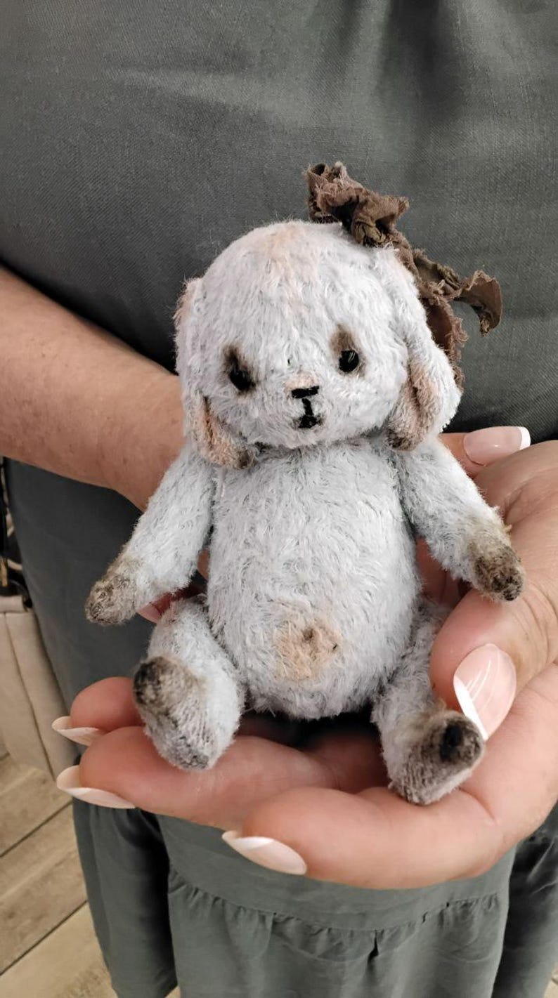 May include: A small, gray and white plush bunny with a brown fabric bow on its head. The bunny is sitting in a person's hand.