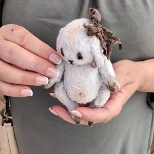 May include: A white handmade teddy bear with brown fabric ears and a brown fabric bow on its head. The bear is sitting in the hands of a person.
