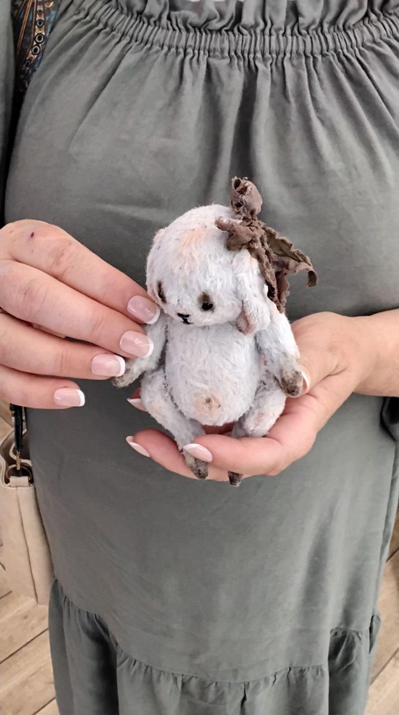 May include: A white handmade teddy bear with brown fabric ears and a brown fabric bow on its head. The bear is sitting in the hands of a person.
