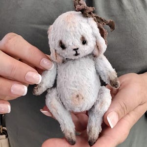 May include: A small, white, handmade plush toy dog with brown accents. The dog has a brown bow on its head and is sitting in a person's hands.