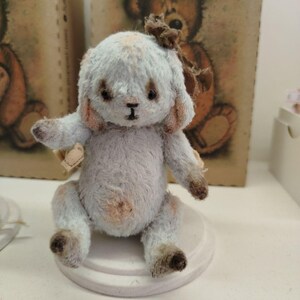 May include: A small, light blue and brown teddy bear with a brown bow. The bear is sitting on a white pedestal.