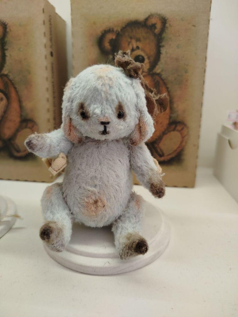 May include: A small, light blue and brown teddy bear with a brown bow. The bear is sitting on a white pedestal.