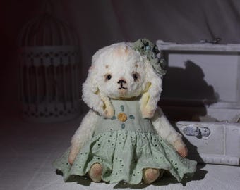 Puede incluir: Un oso de peluche blanco hecho a mano con orejas largas, que lleva un vestido verde con ribete de encaje blanco y una flor verde en la cabeza. El oso está sentado sobre una superficie blanca.