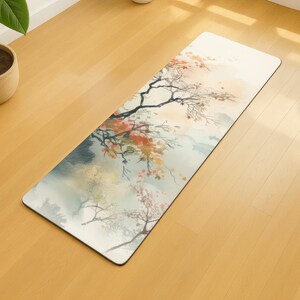 May include: Rectangular yoga mat with a watercolor design of a tree with orange and pink blossoms. The mat has a light background with a mountain scene and is placed on a wooden floor.