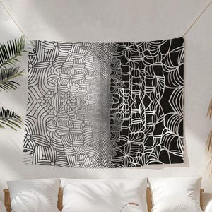 May include: A black and white tapestry with a geometric design hangs on a wall. The design fades from white to black, featuring intricate patterns of lines and shapes. The tapestry is suspended by a string, with a decorative tassel nearby.