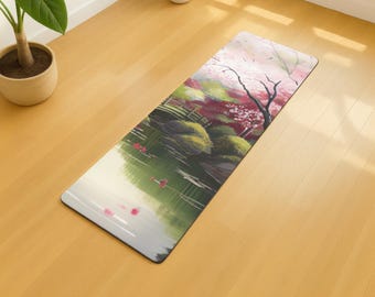 May include: Rectangular yoga mat featuring a serene Japanese garden scene. The design includes a pond with pink flowers, lush green foliage, and a cherry blossom tree. The mat is placed on a light-colored wooden floor.