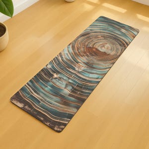 May include: Rectangular yoga mat with a wood grain design. The mat features concentric circles in shades of brown, teal, and white, resembling a cross-section of a tree trunk. The mat is placed on a light-coloured wooden floor.