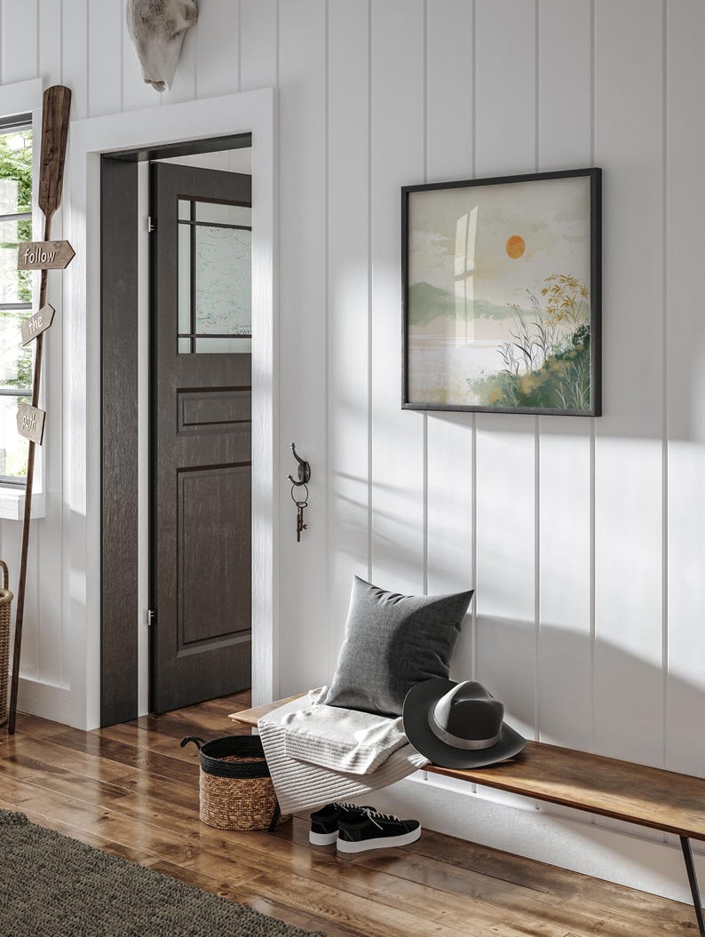 May include: A wooden bench with a gray pillow, a white blanket, a black hat, and a woven basket. The bench is in a hallway with a white wainscoting wall, a dark wood door, and a framed print of a landscape with a sun.