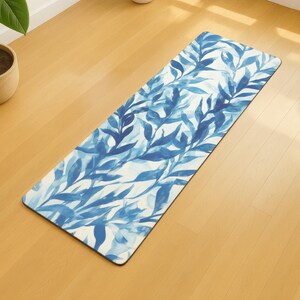 Op de afbeelding: Een rechthoekige yogamat met een blauw en wit aquarel bladpatroon. De mat ligt op een lichtbruine houten vloer. Een plant in een pot is zichtbaar op de achtergrond.