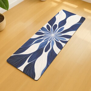 Op de afbeelding: Een rechthoekige yogamat met een bloemmotief in blauw- en wittinten. Het patroon lijkt op een gestileerde bloem met langwerpige bloemblaadjes die uitstralen vanuit een centraal punt. De mat ligt op een licht houten vloer.