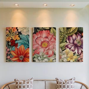 Op de afbeelding: Drie bloemen canvas kunstwerken. Het kunstwerk toont grote, kleurrijke bloemen in tinten oranje, roze, paars en geel tegen een donkere achtergrond. De kunst wordt tentoongesteld op een witte muur boven een bank.