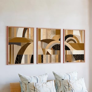May include: Three framed abstract art prints in shades of brown, beige, and black hang on a white wall. The artwork features geometric shapes and curved lines. Below, patterned pillows rest on a wooden bed frame, complementing the art.