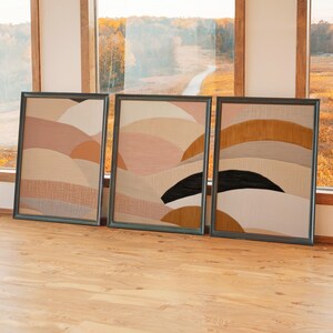 Puede incluir: Tres obras de arte abstractas enmarcadas. Cada una presenta diseños texturizados en capas en tonos neutros de beige, melocotón y marrón, con un acento negro en una. Los marcos son de color gris oscuro y la obra se exhibe sobre un suelo de madera.
