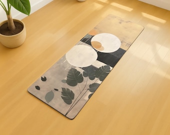 May include: Rectangular yoga mat with a botanical design. The mat features large white circles, dark green leaves, and a color palette of beige, gray, and black. The mat is lying on a light-colored wooden floor.