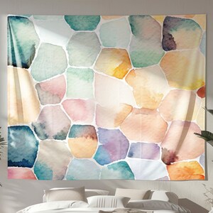 Puede incluir: Un gran tapiz de pared rectangular con un diseño abstracto de acuarela. La obra de arte presenta formas geométricas irregulares en una paleta de colores pastel suaves, incluyendo azules, verdes, rosas y naranjas. El tapiz se exhibe sobre una cama.