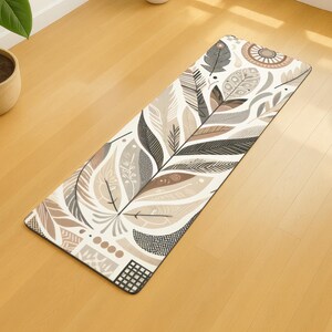 May include: A rectangular yoga mat with a feather design in shades of grey, brown, and cream. The mat has a black border and is on a light-coloured wooden floor. The design includes stylised feathers and geometric shapes.