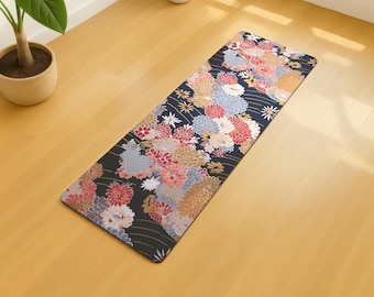 May include: A rectangular yoga mat with a floral pattern in shades of red, pink, blue, and gold against a black background. The mat is placed on a light-colored wooden floor, with a potted plant visible in the background.