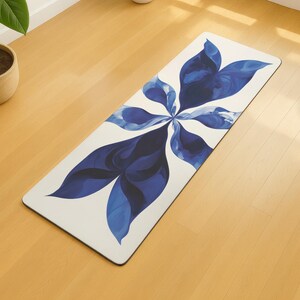 May include: A rectangular yoga mat featuring a white base and a large blue floral design. The floral pattern is a stylized depiction of leaves or petals. The mat is on a light-coloured wooden floor.