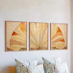 Op de afbeelding: Drie ingelijste abstracte kunstwerken met een waaiervormig ontwerp in tinten geel, oranje en bruin. De kunstwerken zijn aan een witte muur bevestigd boven decoratieve kussens. De lijsten zijn lichtbruin.