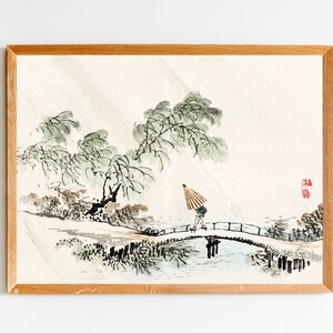 May include: A framed watercolour painting of a person walking across a bridge in a Japanese garden. The painting features a person with an umbrella walking across a bridge over a stream. The background includes trees and a light brown wooden frame.