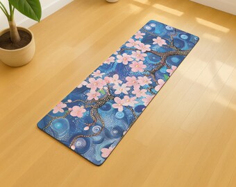 May include: A rectangular yoga mat with a blue background and a floral design. The design features pink cherry blossoms and a tree with brown branches. The mat is made of a durable material and is suitable for yoga and exercise.