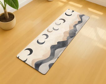 May include: A rectangular yoga mat with a cream background and a design of crescent moons and abstract mountain shapes in grey, beige, and brown. The mat is on a light wooden floor. The mat's design is reminiscent of a landscape.