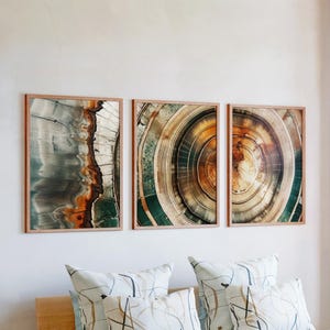 May include: Three framed abstract art prints. The prints feature close-up views of wood grain patterns in shades of gray, green, brown, and orange. The frames are a light brown color. The prints are displayed above a bed with patterned pillows.