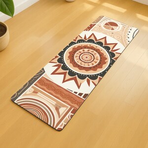 Puede incluir: Una esterilla de yoga rectangular con un diseño geométrico en tonos marrón, negro y crema. La esterilla presenta un patrón circular central con líneas radiales y un borde de varias formas. La esterilla está colocada sobre un suelo de madera.